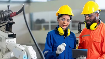 Two engineers in overalls and hard hats look at tablet next to robotic arm 