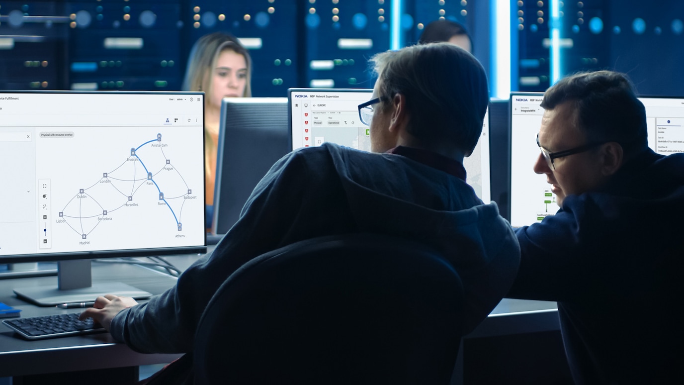 Two men in control room looking at network display on monitors