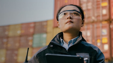 Port employee checking details on tablet device in front of stacked shipping containers