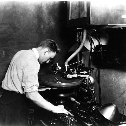 Close-up view of a recording turntable. An attendant is shown viewing the record-cut through a microscope.