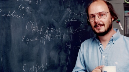 Bjarne Stroustrup standing infront of a chalkboard with a white mug of coffee