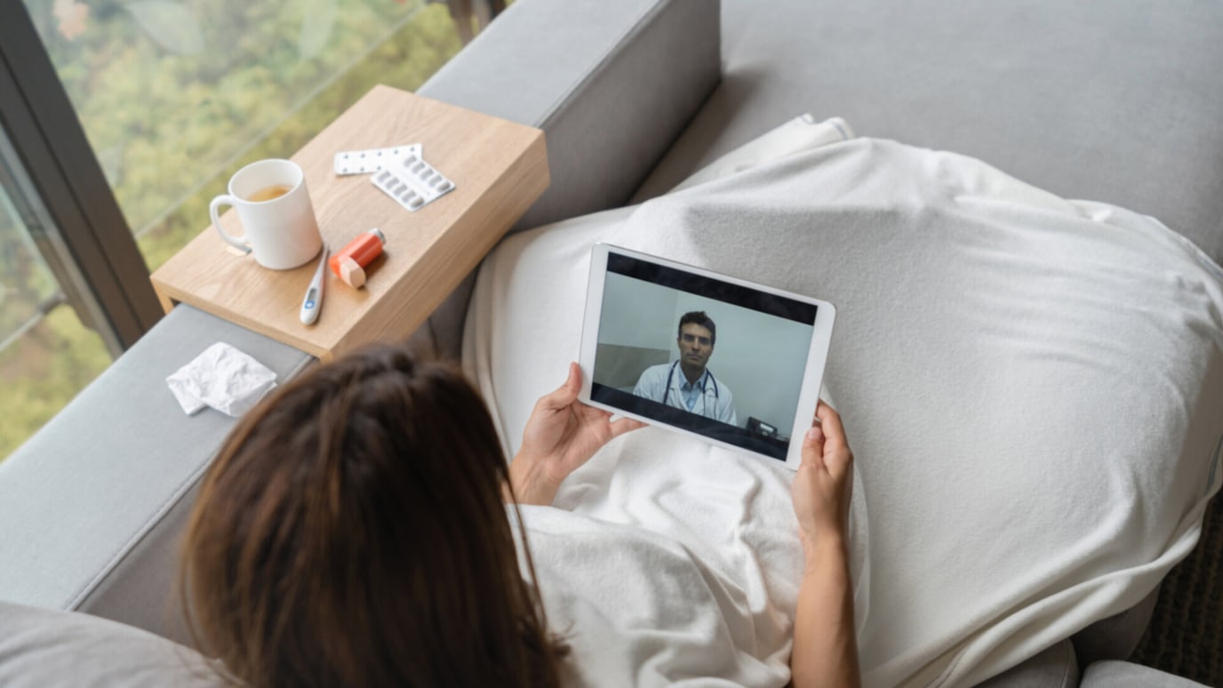 A woman lying on a couch, holding a tablet and having a video call with a doctor. There are medical supplies on a table nearby, suggesting that she is unwell.