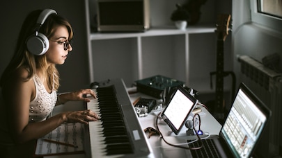 a young woman in a home music studio setting. She's wearing headphones and glasses, seated at a digital piano keyboard.