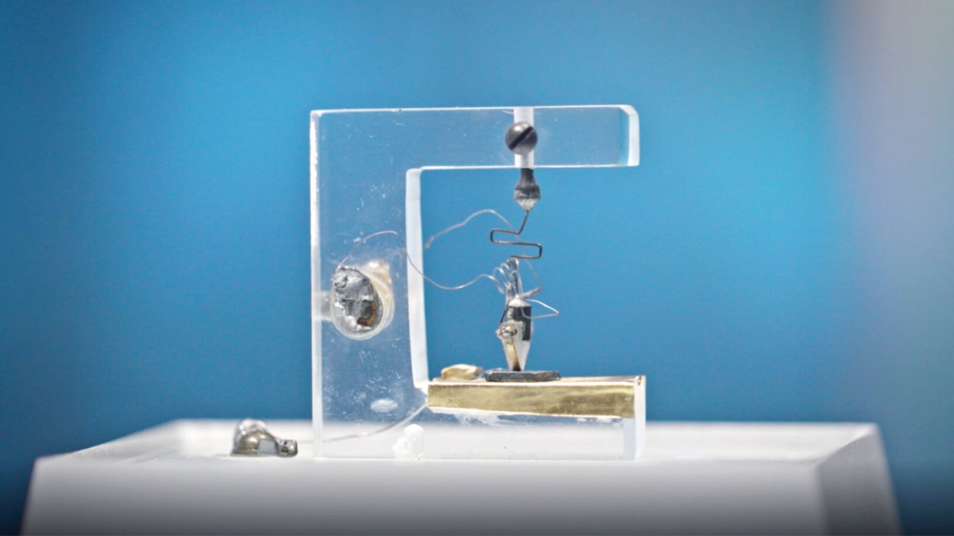 Close-up of the first point-contact transistor invented at Bell Labs in 1947. The device is mounted on a clear plastic stand, showing its small size and intricate wire structure. It sits on a white pedestal against a blue background, highlighting its historical significance in electronics.