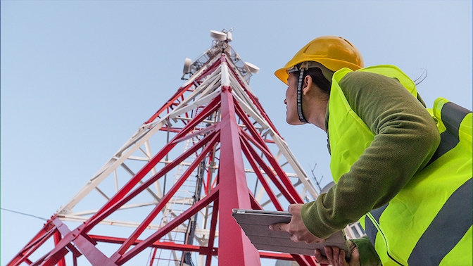 A worker holding a tablet looks up at a communication tower