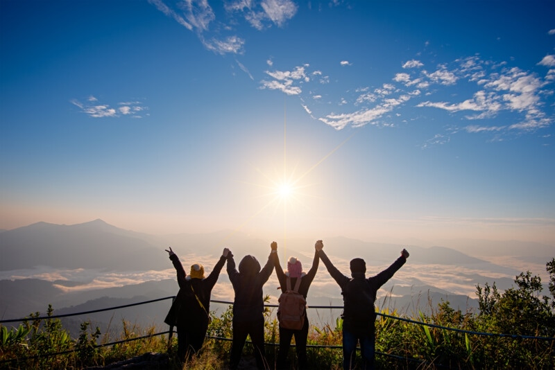four people with hands raised on a mountain looking at the sun