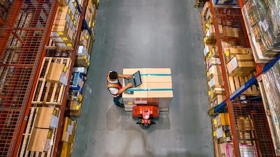 Overhead view of a warehouse worker using a tablet device while standing next to a pallet of boxes. The image is taken from above, showing tall storage racks with cardboard boxes on either side of a concrete aisle.