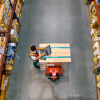 Overhead view of a warehouse worker using a tablet device while standing next to a pallet of boxes. The image is taken from above, showing tall storage racks with cardboard boxes on either side of a concrete aisle.