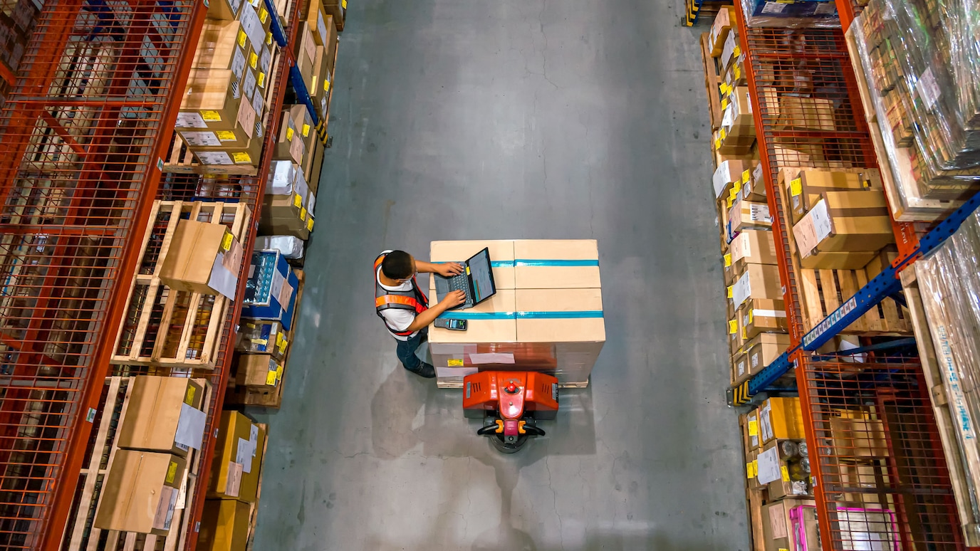 Overhead view of a warehouse worker using a tablet device while standing next to a pallet of boxes. The image is taken from above, showing tall storage racks with cardboard boxes on either side of a concrete aisle.