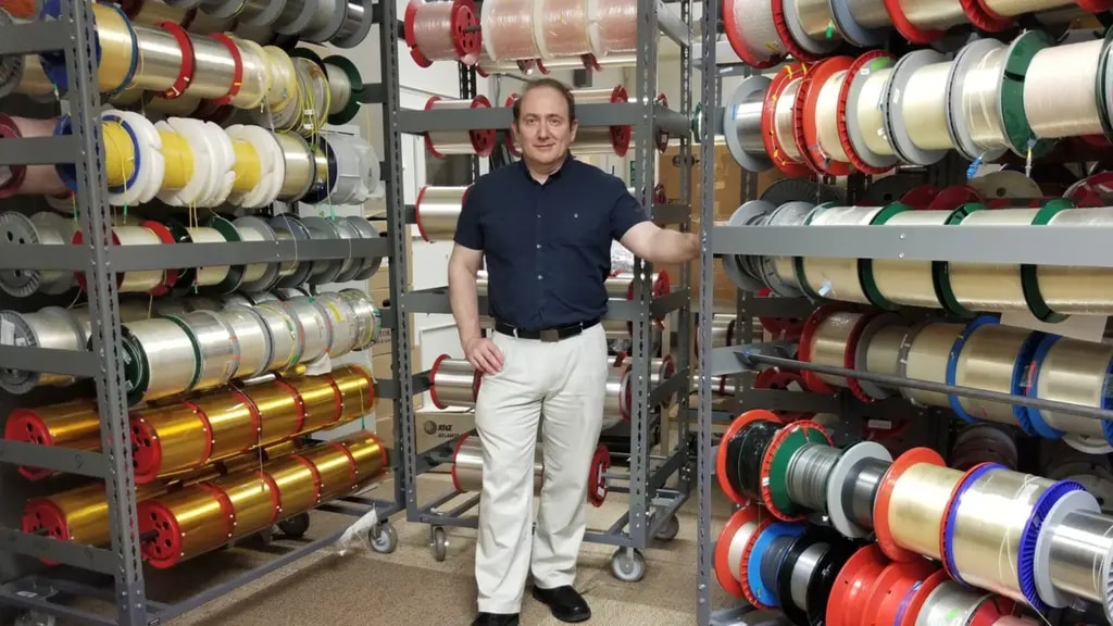 Jean Essiambre standing in a storage room surrounded by shelves filled with large spools of wires or fibers in various colors, including gold, silver, and red.