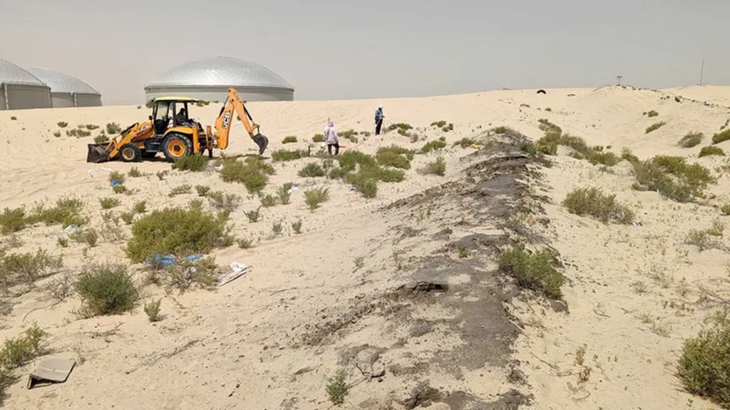 A desert scene with a backhoe loader excavating sandy terrain, accompanied by workers inspecting the area. Sparse vegetation dots the landscape, and dome-shaped storage structures are visible in the background under a hazy sky.