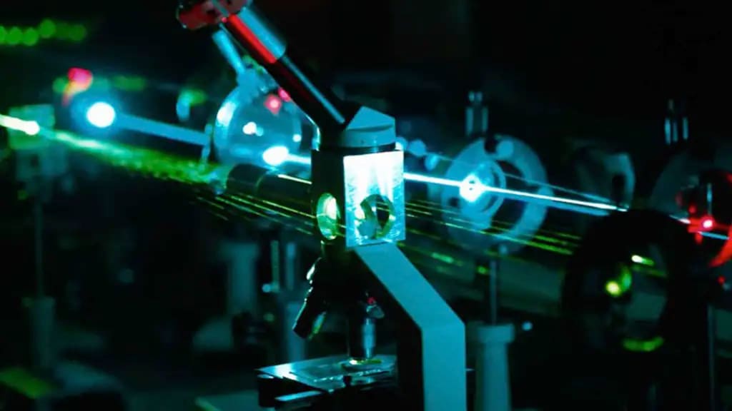 An experimental setup featuring laser beams traveling through optical components, including lenses and mirrors, with bright green and blue light illuminating the dark environment.