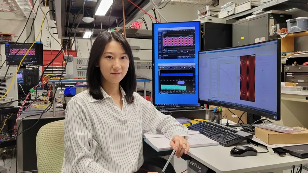 Qian Hu sitting at a desk in the lab, in front of monitors displaying charts and waveforms