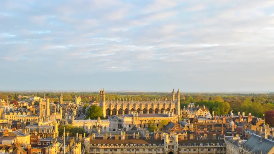 A cityscape of Cambridge, featuring historic buildings and lush greenery under a partly cloudy sky