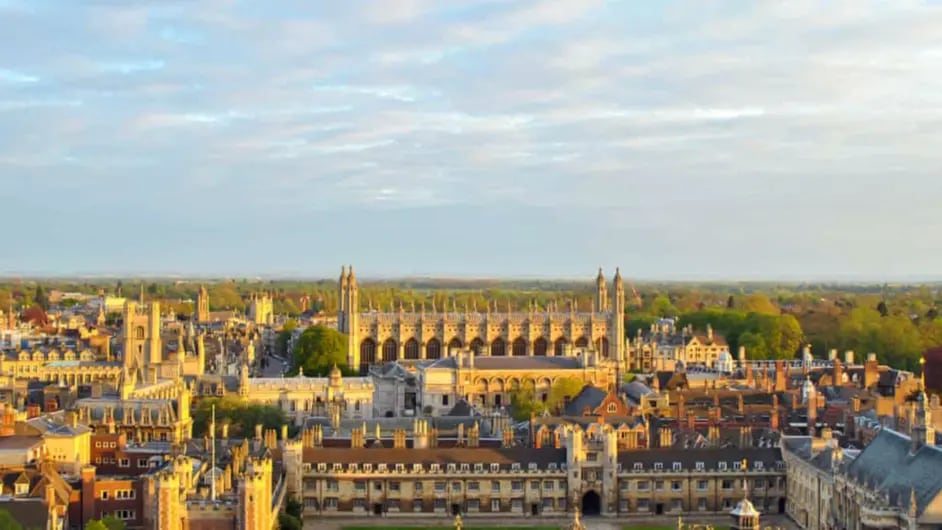 A cityscape of Cambridge, featuring historic buildings and lush greenery under a partly cloudy sky