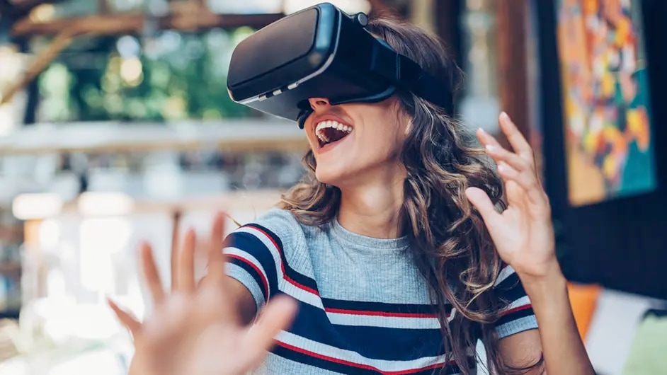 A young woman wearing a VR headset, her hands outstretched and a wide smile on her face. She appears to be immersed in a virtual reality experience.