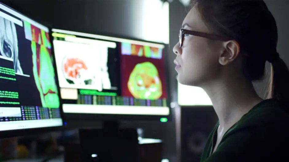 A person working in a dimly lit room, focused on multiple computer screens displaying medical imaging data. The screens show colorful brain scans and other diagnostic images.