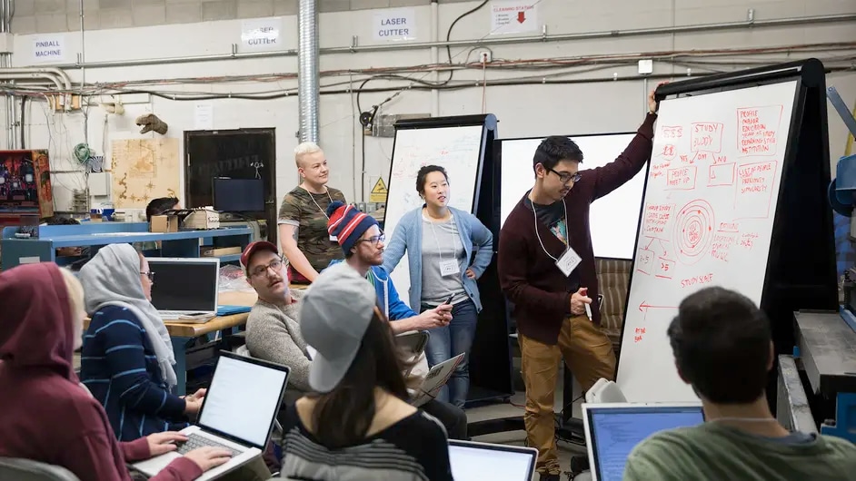 A group of people gathered around a whiteboard in a workshop setting. One person is presenting a diagram about a study buddy system, while others listen and take notes on their laptops.