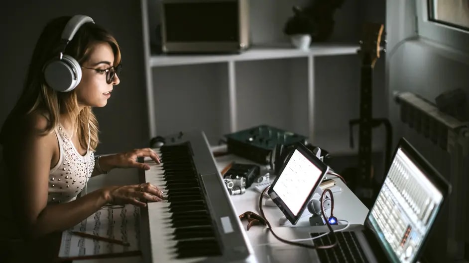 a young woman in a home music studio setting. She's wearing headphones and glasses, seated at a digital piano keyboard.