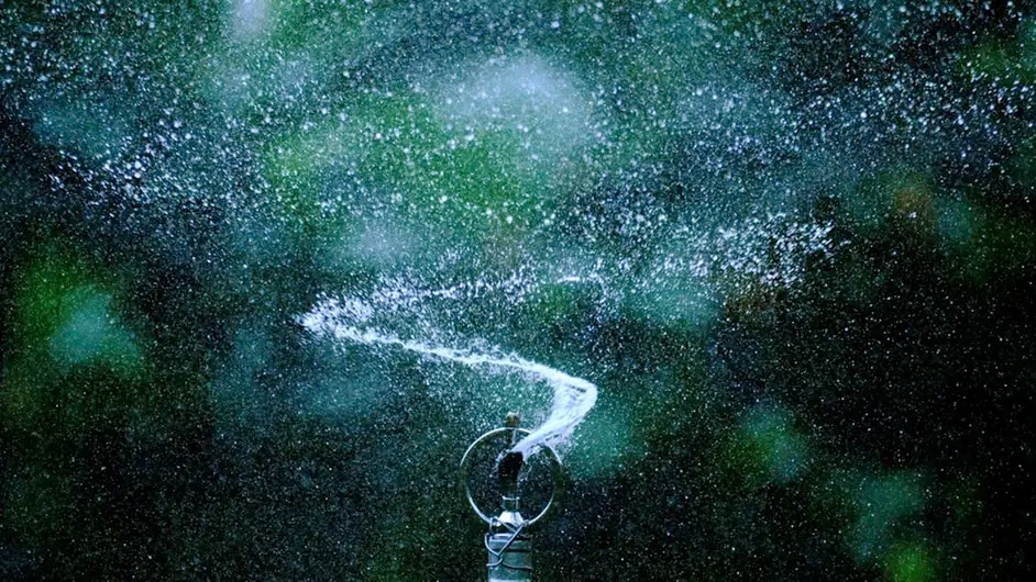 A close-up of a sprinkler spraying water in a circular motion against a green background